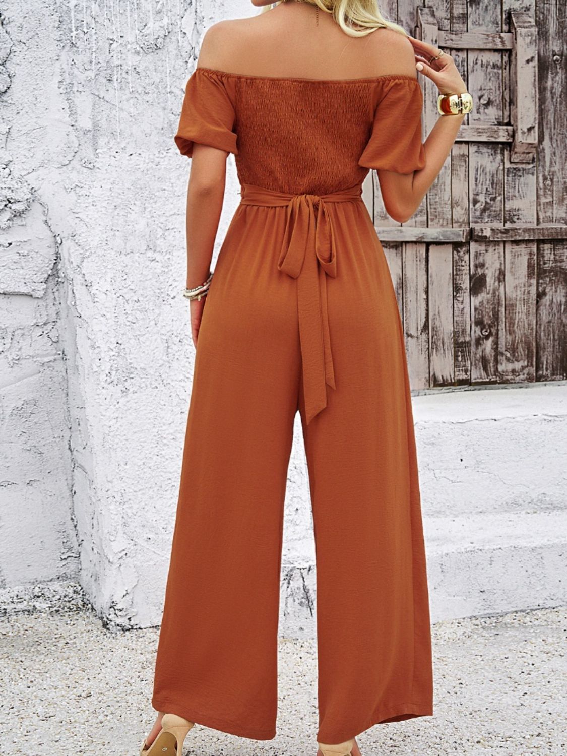 Rust-colored off-shoulder jumpsuit with wide-leg pants and a waist tie.